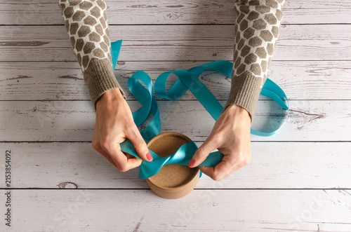 Woman preparing a gift box with a blue Ribbon on white wooden table.