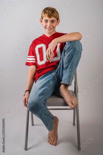 Smiling barefoot boy wearing red American football jersey and jeans sitting with one leg up on a folding chair