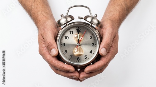 Hands holding a classic alarm clock with hands nearing the hour, symbolizing the passage of time and the urgency of deadlines. The metallic surface of the clock reflects light.