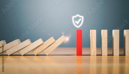 Red wooden block standing as a protective barrier against falling dominoes, with a glowing shield above, representing risk management, security, and business continuity.