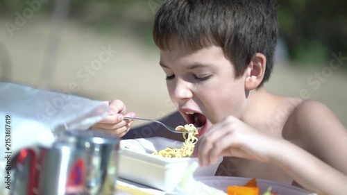 Shirtless boy eats instant noodles with a fork from a styrofoam box at an outdoor table on a sunny beach, focused and enjoying a simple meal in nature.