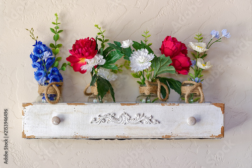 Silk flowers of various colors are displayed on the wall on a shelf made from an antique drawer