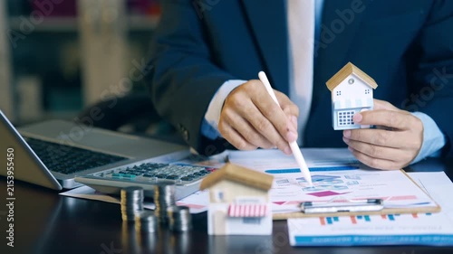 Real estate agent holding house model and using pen to evaluate property price and offer details for clients.