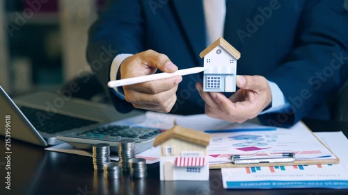 Real estate agent holding house model and using pen to evaluate property price and offer details for clients.