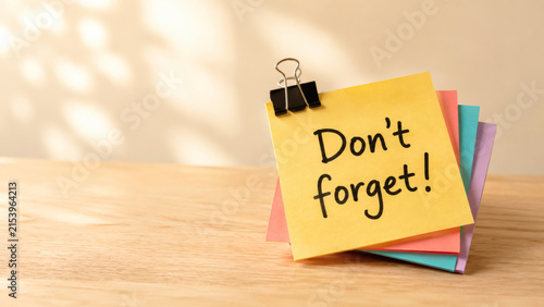 Don't Forget: A sticky note with the reminder Don't forget! held by a binder clip, placed on a wooden desk surface bathed in soft, dappled sunlight.