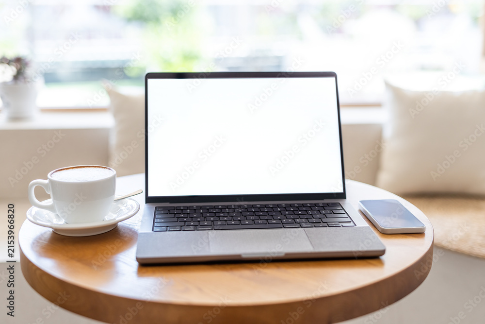 Fototapeta premium A blank screen laptop, coffee cup and smartphone placed on a round wooden table in a bright indoor space.