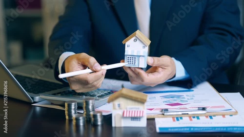 Real estate agent holding house model and using pen to evaluate property price and offer details for clients.