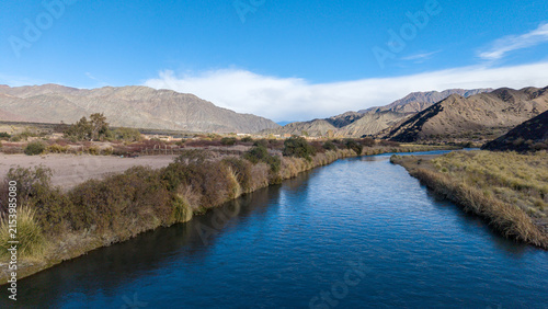 Aerial view of the San Juan River near the abandoned aluminum sulfate plant ruins in Calingasta, San Juan, Argentina, surrounded by mountains and shrubs under a clear blue sky.