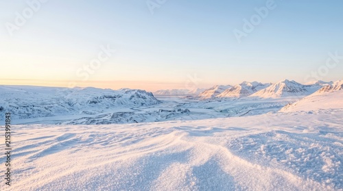 A wide, panoramic view of a snowy, mountainous landscape at sunrise. The foreground is covered in fresh snow, with visible tracks in the snow.