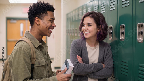 Two happy high school students laughing and talking together while standing next to green lockers in a school hallway.
