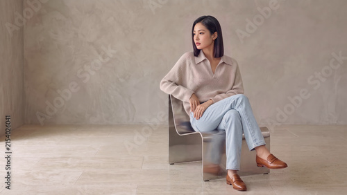 A female model in a beige sweater, light blue jeans, and brown loafers sits on a modern metal chair in a minimalist setting, looking left across expansive negative space.