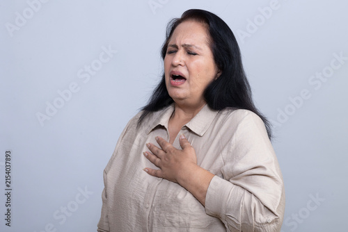 Canvastavla Mature Indian woman holding her chest while experiencing apparent chest pain and discomfort, showing a distressed expression in a clean studio setting, representing a health and medical concern