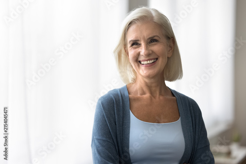 Happy retirement time. Cheerful beautiful older age blond woman stand indoors look aside with pretty smile. Joyful active aged female pensioner feel well healthy laugh enjoy positive moments in life