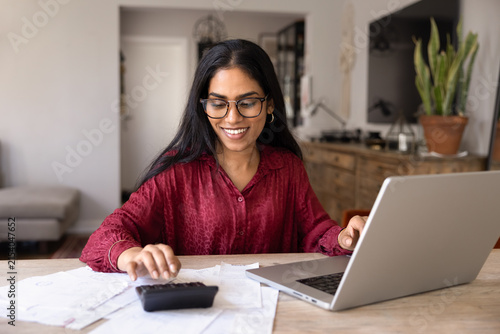 Canvas Print Smiling Indian woman with glasses using calculator and online banking on laptop
