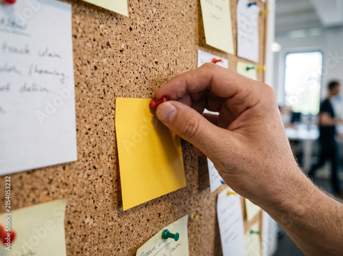 Hand pinning bright yellow sticky note on cork notice board in office