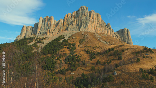 Bild auf Leinwand Dramatic rocky mountain peak bathed in warm golden light with evergreen trees dry grass on slopes