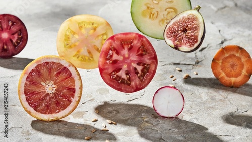 Assorted fresh fruit and vegetable slices floating above a textured grey stone surface with scattered seeds and natural lighting conditions