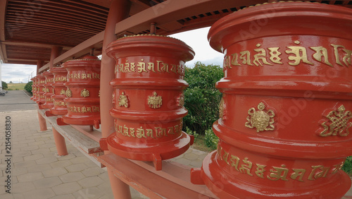 Obraz na plátně Row of vibrant red prayer wheels with gold Tibetan script decorative symbols under wooden shelter