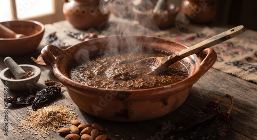A rustic, atmospheric photograph of mole poblano simmering slowly in a traditional red earthenware clay pot (cazuela de barro), with steam rising and toasted seeds