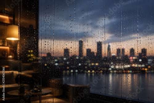 Rainy evening view of a city skyline from a cozy indoor lounge with water droplets on the window.