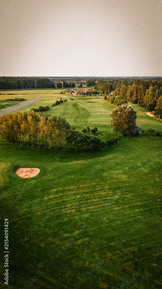 Fototapeta premium Aerial view of a lush green golf course with manicured fairways, sand traps, and trees under a clear sky, showcasing the serene landscape of a recreational area