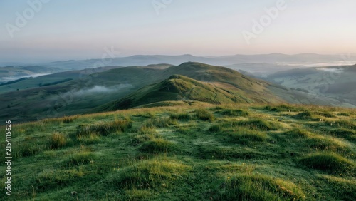 Rolling green hills covered in lush grass under a soft morning sky with patches of mist settling in the valleys of a peaceful rural landscape