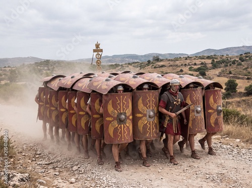 Roman legionaries in testudo formation march in dusty landscape, ancient military tactic, historical reenactment