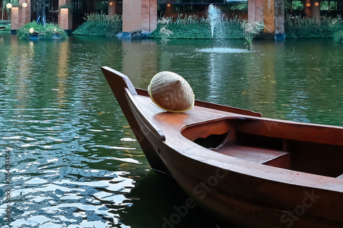 A traditional wooden boat with a woven conical hat resting on its bow floats calmly on a scenic lake featuring a distant water fountain.