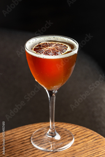 Cocktail in a stemmed glass with a dried orange slice garnish on a wooden table against a dark background, showcasing rich amber color and bubbles