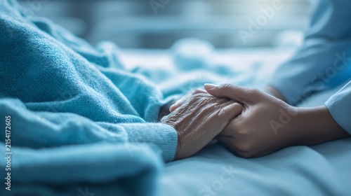 young healthcare worker hands holding the hand of an elderly patient resting on a hospital bed. Soft blue blanket texture visible in the background. Calm and peaceful atmosphere representing 