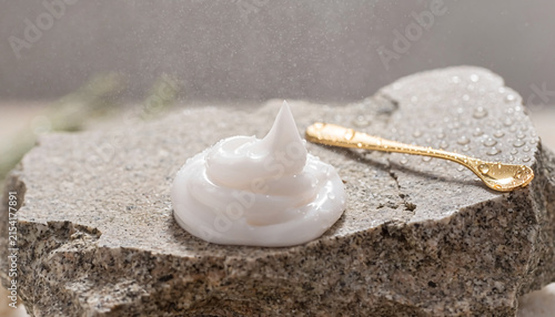 White Facial Cream Swirl and Gold Cosmetic Spoon on Natural Stone Podium with Water Droplets