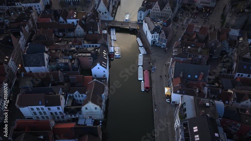 Cinematic aerial Ghent Belgium reveals medieval architecture lining waterways with moored tour boats. Historic gabled houses and bridges frame the canal as evening light bathes the Flemish cityscape.
