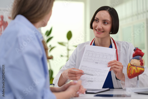 Woman doctor shows cardiogram sheet to lady patient near heart model in clinic. Smiling cardiologist explains test result while points to ECG lines