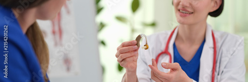 Woman doctor shows hearing aid during audiology consultation in clinic. Smiling specialist presents small device explaining to patient sound support