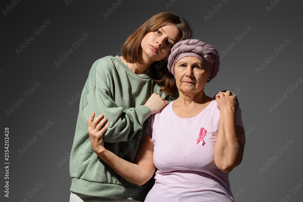 Fototapeta premium Senior woman after chemotherapy with pink ribbon and her granddaughter hugging on dark background. Breast cancer awareness concept