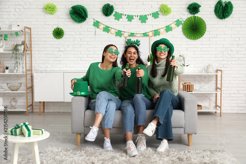 Beautiful young women with bottles of beer on sofa at home. St. Patricks Day ...