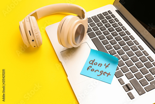 Sticky note with text DONT FORGET, laptop and headphones on yellow background, closeup