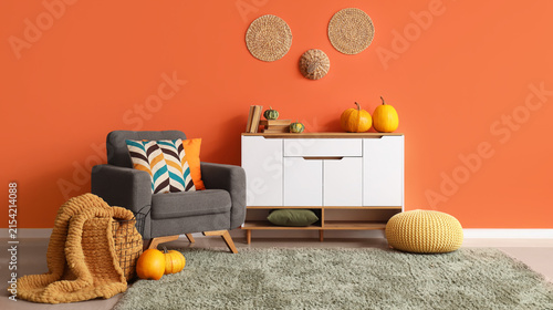 Cozy autumn living room interior with gray armchair  white cabinet  and pumpkins against an orange wall