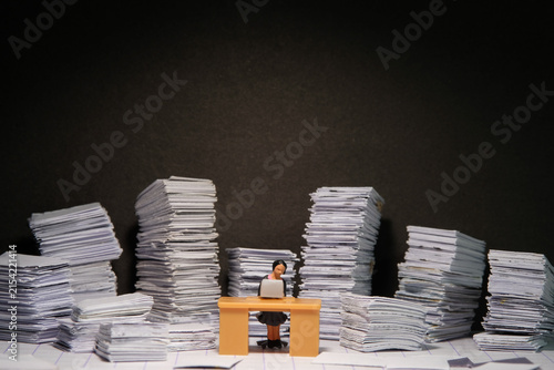 Miniature tiny people toy figure photography. Overload and work overtime concept. A female businesswomen seat on a desk with pile of document, working until night at dark room with spotlight