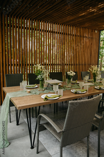 Interior of a banquet hall made of wood in the form of a gazebo in an open space with dishes, food, drinks and flowers in vases and a green tablecloth on the table for a wedding or anniversary 