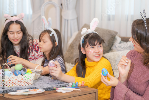 Konstfotografi At a wooden coffee table strewn with brushes, paints and eggs, a modern multicultural family of a mother and three excited young daughters raising a yellow painted egg beside a smiling face