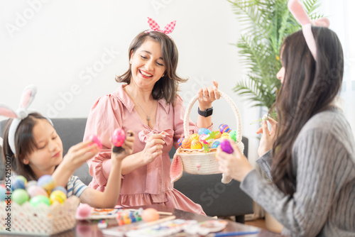 Fotografie A mother and her two daughters passing a freshly painted egg from one hand to another at a wooden coffee table strewn with brushes, paints and eggs on a happy holiday morning