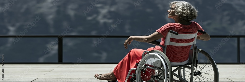 Fototapeta premium Senior woman in wheelchair seated on terrace with dramatic mountain backdrop for accessibility poster and outdoor wellness design