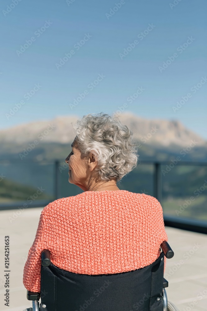 Fototapeta premium Rear view of elderly woman in wheelchair on balcony with distant mountain vista for senior care banner and peaceful background design