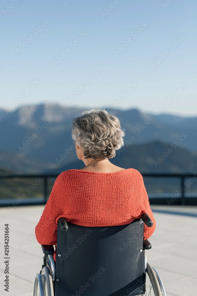 Fototapeta premium Rear view of an elderly woman in a wheelchair facing mountain vista on a balcony for serene background and healthcare design use