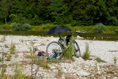 A bicycle with a mounted umbrella is parked on the riverbank of the renaturalized Isar at Flaucher near Munich. Creative sun protection for a sunbather
