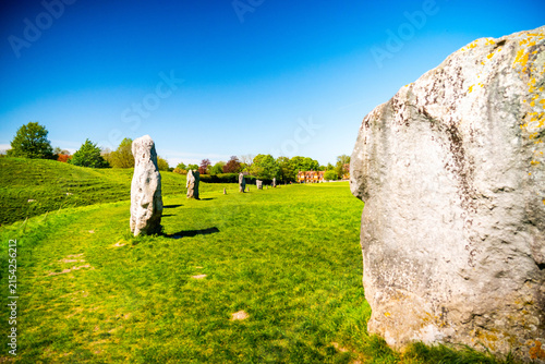 View of the ancient Neolithic standing stones of Avebury Stone Circle set in a lush green meadow under a clear blue sky in Avebury, Wiltshire, United Kingdom.