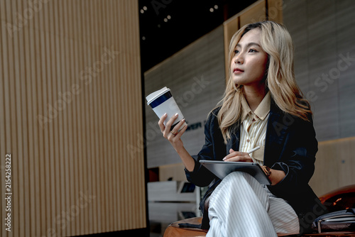 Fotografía Asian businesswoman lifts her coffee cup toward her lips while glancing sideways, tablet resting on her lap in a warmly lit wood-paneled lobby in Bangkok's business district