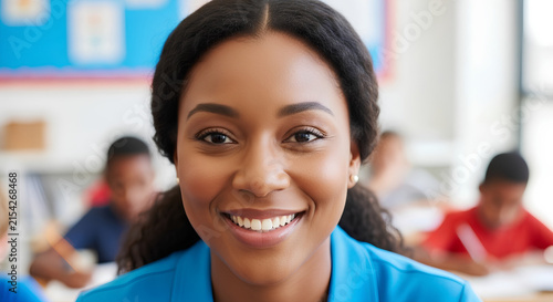 A smiling female teacher in a blue shirt looks directly at the camera in a classroom with students in the background.