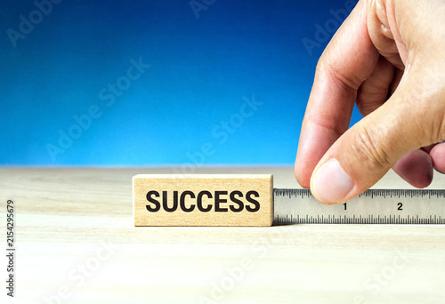 Human hand measuring a wooden block labeled success with a ruler on table